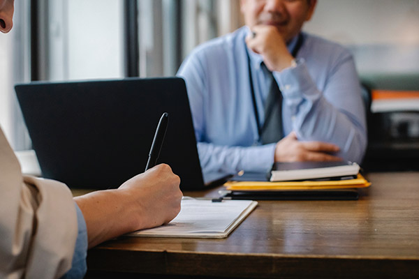 Someone signs a document across the table from a man in a button-up shirt.